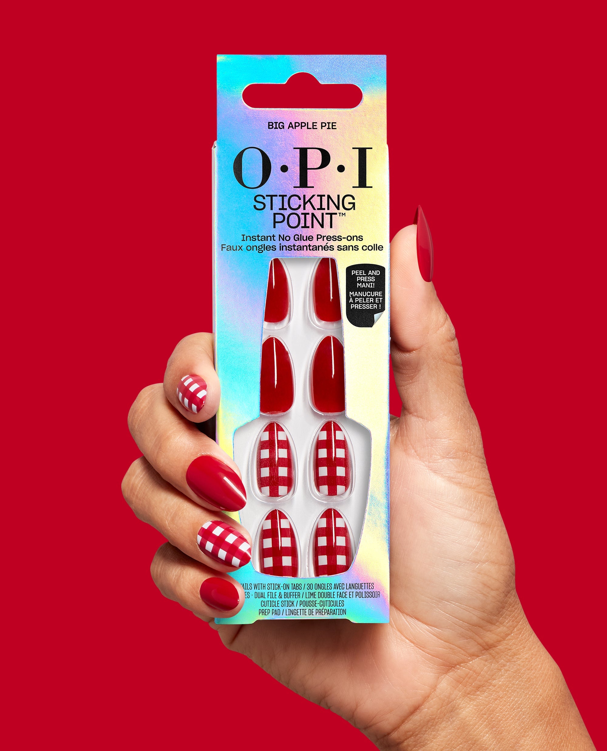 Hand holds OPI Sticking Point Big Apple Pie instant no-glue press-on nails, featuring glossy red and red-white gingham designs, with matching manicure on a bold red background for a trending nail look.