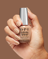 Hand with neatly manicured nude-beige nails holds an OPI nail polish bottle labeled A Total Suzi, Infinite Shine gel-like lacquer, against a warm brown background, showcasing the glossy neutral shade for US shoppers.