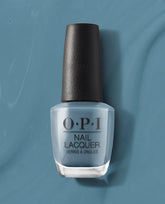 OPI Alpaca My Bags Nail Lacquer bottle showcasing a muted dusty blue with gray undertones, standing upright against a coordinating teal-blue background, perfect for cool-toned fall nails.