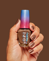 Hand with glossy brown nails holds OPI RapiDry Awe of a Sudden nail polish, a rich shimmer brown fall nail color, with a blue-to-pink cap on a warm brown background.