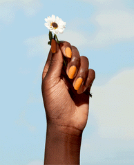 Manicured hand with honey-yellow nails holds a white daisy against a blue sky, showcasing OPI Nature Strong Vegan Nail Lacquer, Bee the Change - a bright, eco-friendly pick for spring nail colors and spring nail trends.