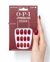 OPI Big Apple Red xPRESS/ON press-on nails in glossy classic red shown in retail packaging beside a hand wearing matching gel-like manicure nails on a white background.
