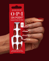 OPI On Point Big Candy Apple Red press-on nails shown on a hand holding the matching red package, with glossy deep red fall nail colors and gold dot accents against a burgundy background.