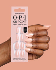 Hand wearing pale pink almond press-on nails holds OPI On Point Bubbly Behavior Instant Press-On Mani packaging, a soft neutral option for fall nail colors on a peach background.