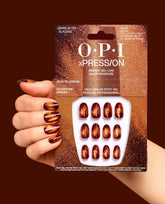 OPI xPRESS/ON Burn After Glazing press-on nails are held beside a matching manicure, showing short court gel-like nails in shimmering red, copper, and amber fall nail colors on a dark brown background.