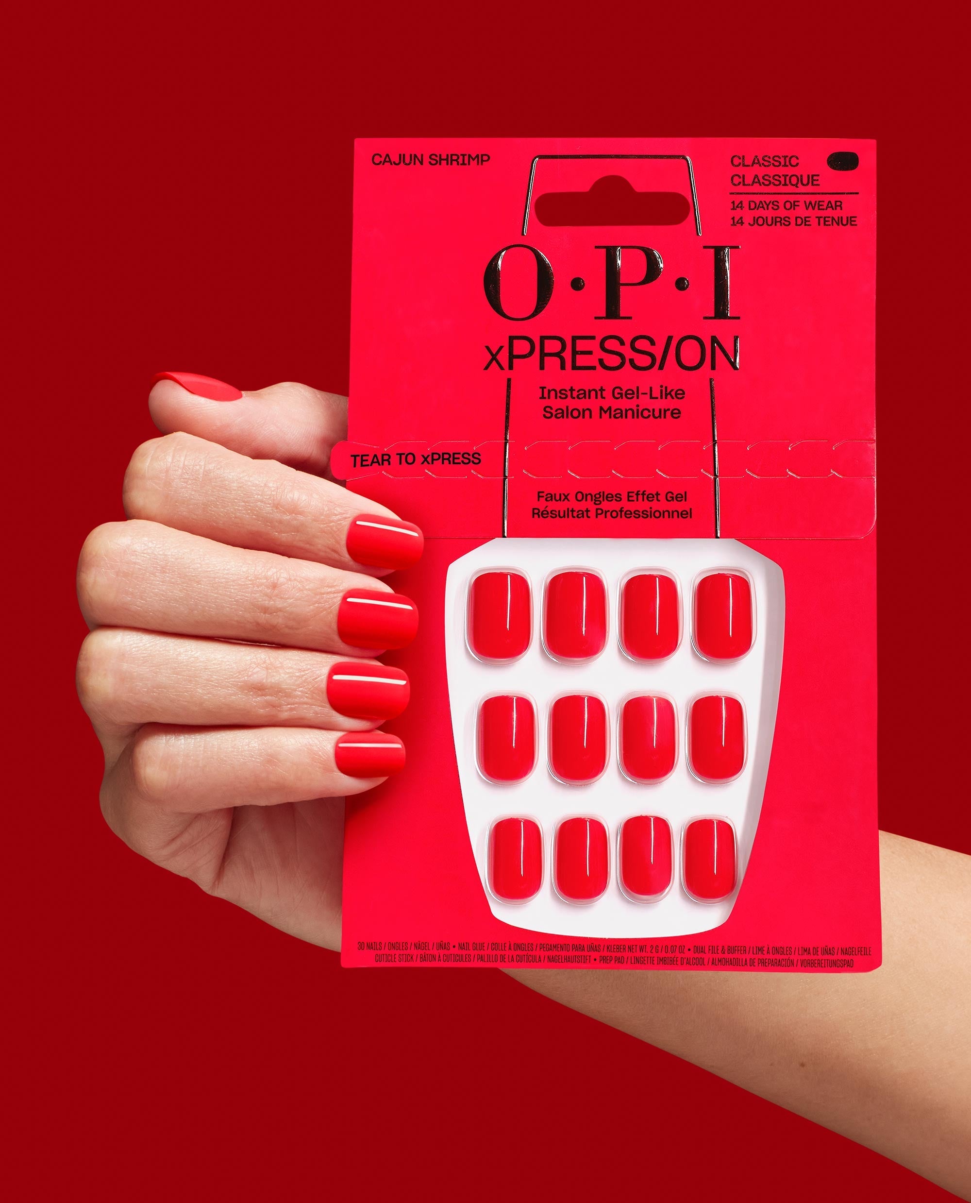 Hand with glossy red OPI xPRESS/ON Cajun Shrimp press-on nails holds matching instant gel-like salon manicure packaging against a bright red background.