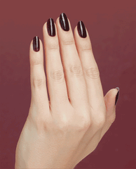 Hand with long oval nails painted in Complimentary Wine, a deep burgundy OPI nail polish nail lacquer, flexing to show glossy, even coverage against a coordinating wine-red background.