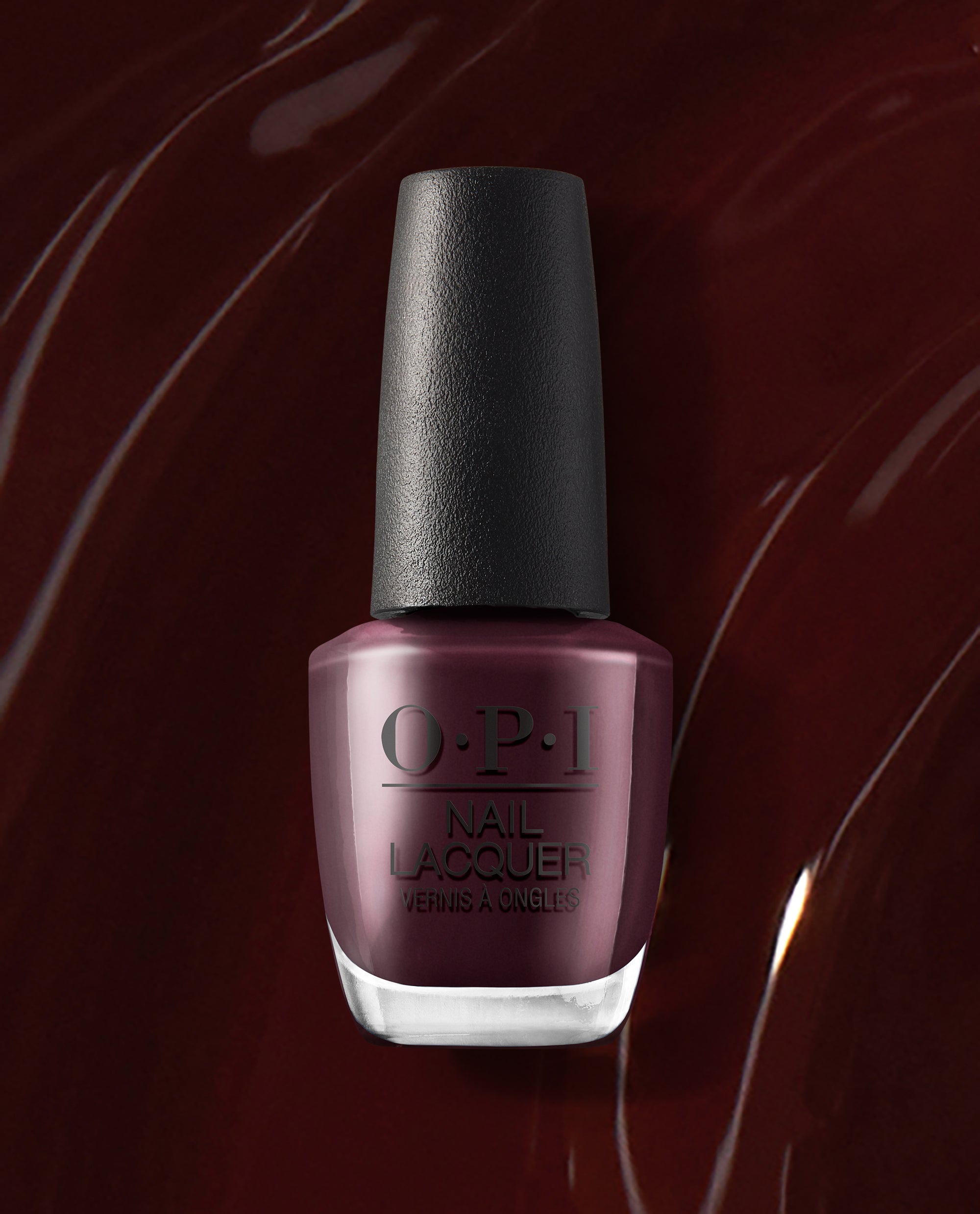 OPI nail polish bottle of Complimentary Wine nail lacquer standing upright, showcasing a deep burgundy-wine shade with a glossy finish against a matching dark red, swirled polish background.