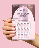 Hand holds OPI xPRESS/ON Don’t Forget to Tip press-on nails in pale pink with metallic taupe French tips, shown in retail packaging against a pink background for chic fall nail colors.