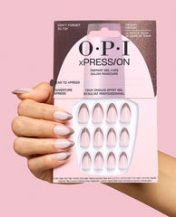 Hand holds OPI xPRESS/ON Don’t Forget to Tip press-on nails in pale pink with metallic taupe French tips, shown in retail packaging against a pink background for chic fall nail colors.