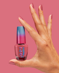 Hand with glossy pink manicured nails holding OPI Dry and Dash RapiDry nail polish bottle, featuring a pink shade and blue-to-pink gradient cap, against a solid pink background, ideal for an OPI nail polish gift set product page.