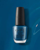 OPI nail polish bottle of Duomo Days, Isola Nights nail lacquer standing upright, showcasing a rich deep teal-blue shade with a black textured cap against a glossy matching blue background.