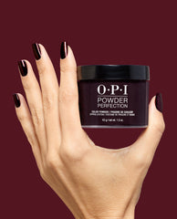Hand with glossy deep burgundy shimmer nails holds OPI Powder Perfection Every Month is Oktoberfest dipping powder, a rich choice for fall nail colors, against a burgundy background.