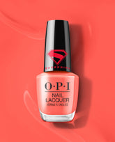 OPI Heat Vision Nail Lacquer bottle stands upright on a glossy coral-red background, showing a peachy coral polish shade and black Supergirl logo cap for a trending manicure look.