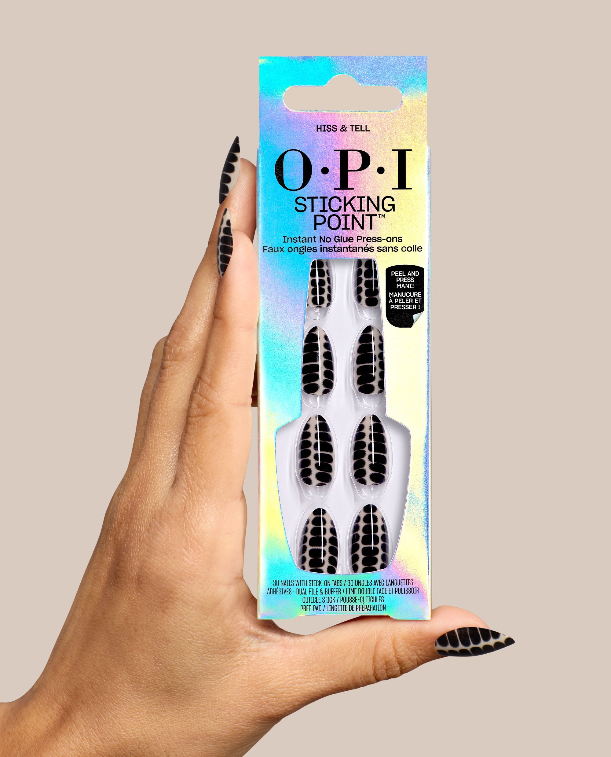 Hand holding OPI Hiss & Tell Sticking Point no-glue press-on nails, featuring black and nude snake-pattern designs in iridescent packaging, styled as bold fall nail colors on a beige background.