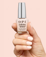 Hand with glossy pale nude manicure holds an OPI Infinite Shine bottle in I’m the Bossa Nova Now, a gel-like lacquer shade for fall nail colors, against a soft peach background.