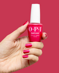Hand with glossy fuchsia nails holds an OPI GelColor It’s All Greek to Me gel nail polish bottle against a matching pink background, showcasing a bright option for fall nail colors.
