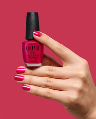 Hand with glossy magenta nails holds OPI It’s All Greek to Me Nail Lacquer, a shimmering pink shade for bold fall nail colors, against a matching pink background.