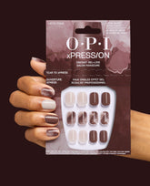 OPI xPRESS/ON Latte-tude press-on nails are held in packaging beside a manicured hand, showing extra-short beige, mocha brown, and marbled gel-like fall nail colors on a dark burgundy background.