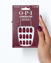 OPI Malaga Wine xPRESS/ON press-on nails in deep burgundy are displayed in classic packaging, held beside a matching manicure for rich fall nail colors.