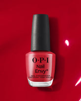 OPI Nail Envy Big Apple Red nail strengthener bottle with black cap stands against a glossy red polish background, showcasing a bold red treatment shade for fall nail colors.