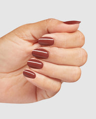 Hand displaying glossy reddish-brown OPI Infinite Shine Nail Prep Rally polish on short rounded nails, a warm shade among fall nail colors on a clean light gray background.