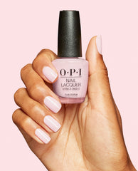 Hand with glossy pale pink manicured nails holds an OPI Nail Lacquer bottle in OPI’m a Bubble Bunny, a soft pastel pink shade, against a light pink background.