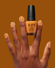 Hand with neatly manicured nails painted in Penny Loafer Lane, a warm caramel OPI nail polish nail lacquer, holding the matching bottle against a solid burnt orange background, showcasing the rich fall nail color.