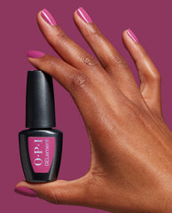 Hand with glossy magenta nails holds an OPI GelColor bottle in Pompeii Me in Nails, an at-home gel nail polish shade for bold fall nail colors, against a matching berry background.