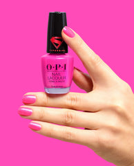 Hand with glossy hot pink nails holds OPI Power up Pink Nail Lacquer, a shimmering pink polish bottle with a black Supergirl cap, against a bright pink background.