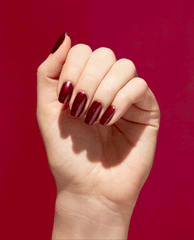 Hand showing glossy deep raisin-red nails painted with OPI Nature Strong Vegan Nail Lacquer in Raisin Your Voice, a rich shimmer shade for fall nail colors, against a red background.