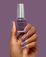 Hand with glossy mauve-purple nails holds OPI Infinite Shine Reign or Shine gel-like lacquer, a muted purple shade for fall nail colors, against a matching purple background.