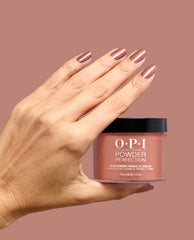 Hand holding OPI Powder Perfection Route Beer Float dipping powder, with nails painted a shimmery warm rose-brown shade against a mauve background for fall nail colors.