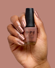 Hand with shimmery rose-brown nails holding OPI Route Beer Float nail lacquer bottle, a warm metallic shade for fall nail colors, against a muted mauve background.