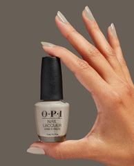 Hand with almond-shaped nails painted in Suzi’s Pager, a taupe-beige OPI nail polish, gracefully holding the matching OPI Nail Lacquer bottle against a solid neutral brown background, showcasing glossy neutral manicure color.
