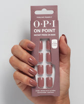 Hand with mauve-pink manicured nails holds OPI On Point Tickle My France-y instant press-on mani package, showing 24 glossy nude press-on nails from an OPI nail polish gift set against a light gray background.