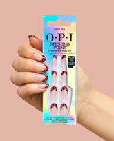 Hand with OPI Tortie Tips press-on nails holds a holographic Sticking Point box, showing nude nails with tortoiseshell French tips for chic fall nail colors on a peach background.