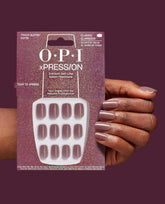 Hand wearing OPI xPRESS/ON Track Suited Cutie mauve shimmer press-on nails holds the matching 30-nail instant gel-like salon manicure pack against a deep plum background, showcasing fall nail colors.