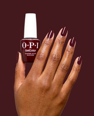 Hand holds OPI GelColor Vampire State Building gel nail polish, with glossy deep burgundy fall nail colors on manicured nails against a matching wine-red background.