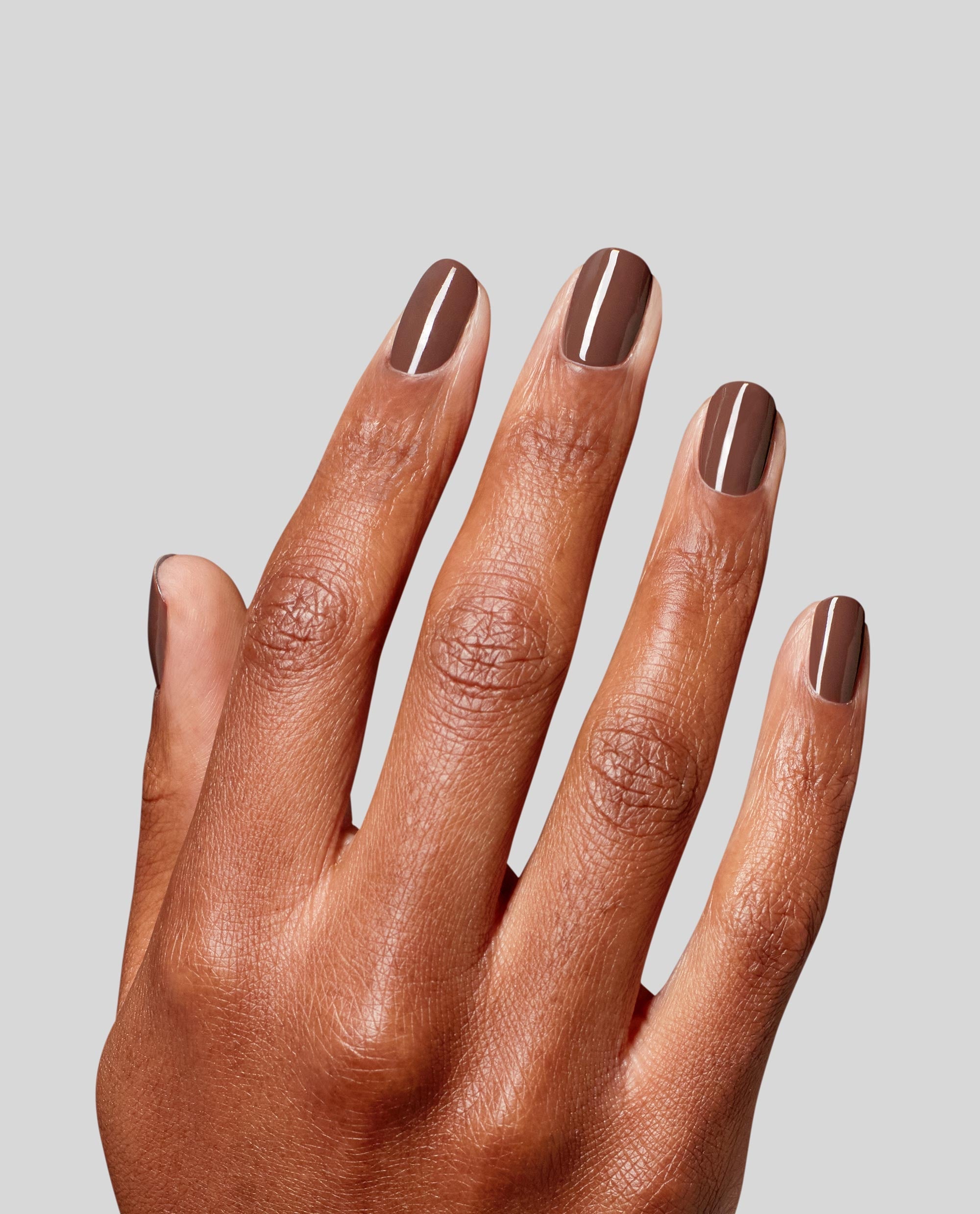 Hand modeling glossy taupe-brown OPI RapiDry nail polish in Impromptu You on short rounded nails, showcasing a chic neutral shade for fall nail colors against a light gray background.