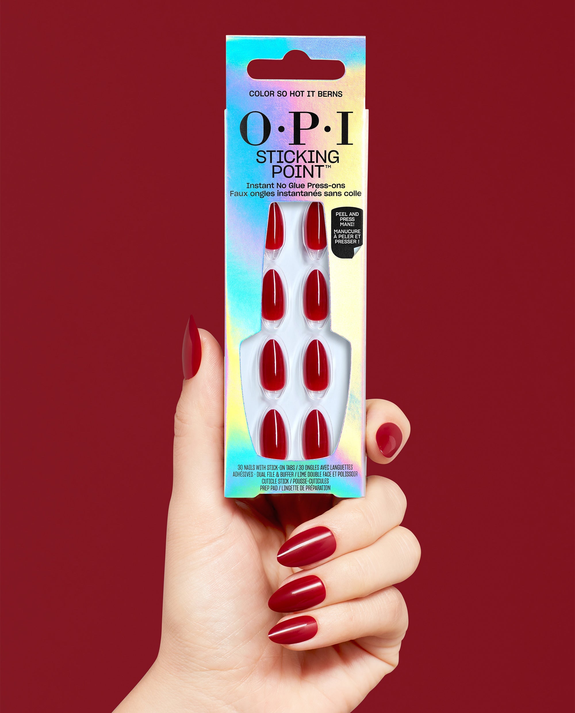 Hand with glossy red almond nails holds OPI Sticking Point Color So Hot it Berns no-glue press-on nails in iridescent packaging against a deep red background.