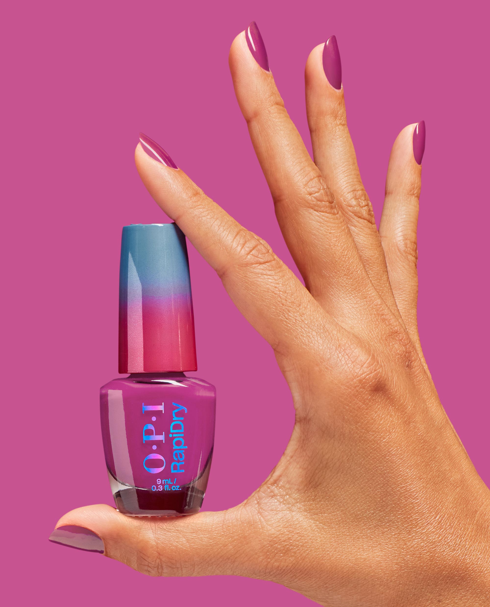 Hand with glossy magenta nails holding an ASAPink OPI RapiDry nail polish bottle against a matching pink background, showcasing the gradient blue-to-pink cap and stylish manicure, ideal for an OPI nail polish gift set product page.