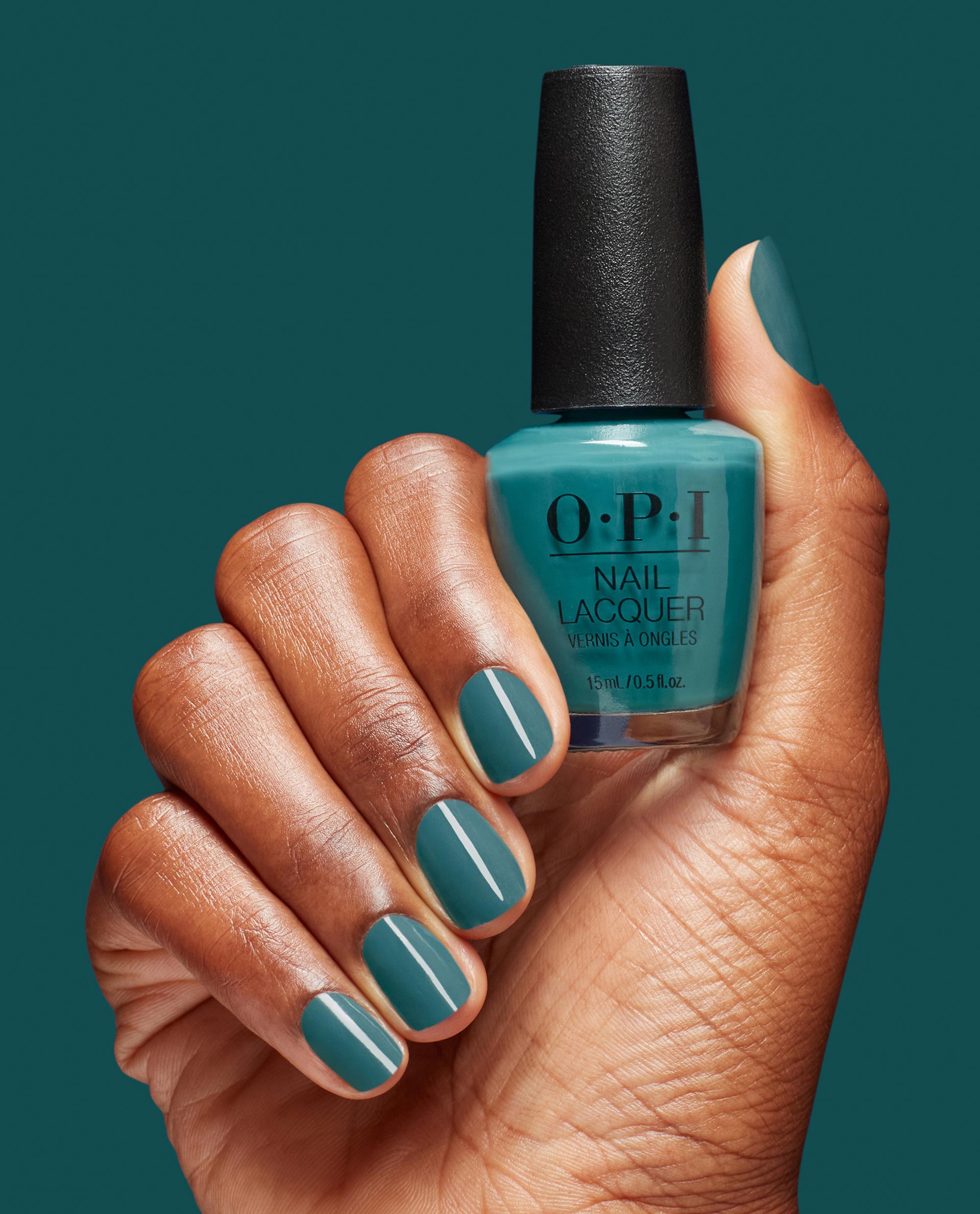 Hand with neatly manicured teal nails holding OPI nail polish Baby Tee-l nail lacquer bottle against a matching teal background, showcasing the glossy finish and modern blue-green shade for US beauty shoppers.