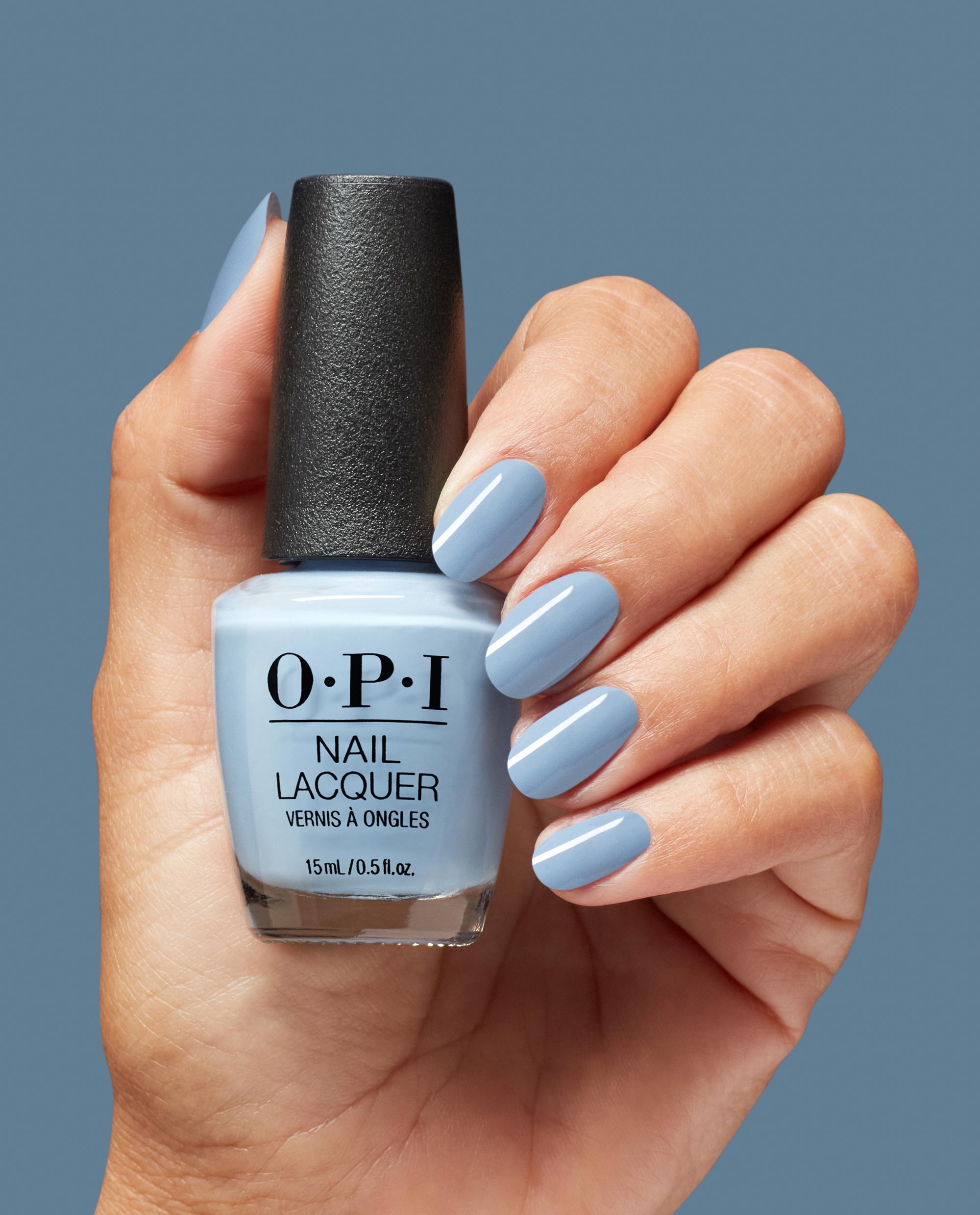 OPI Baggy Jean Baby Nail Lacquer coats oval nails as a hand holds the 15 mL (0.5 fl oz) bottle, showcasing a glossy light denim blue shade against a matching blue backdrop - an on-trend pick for fall nail colors in the US.