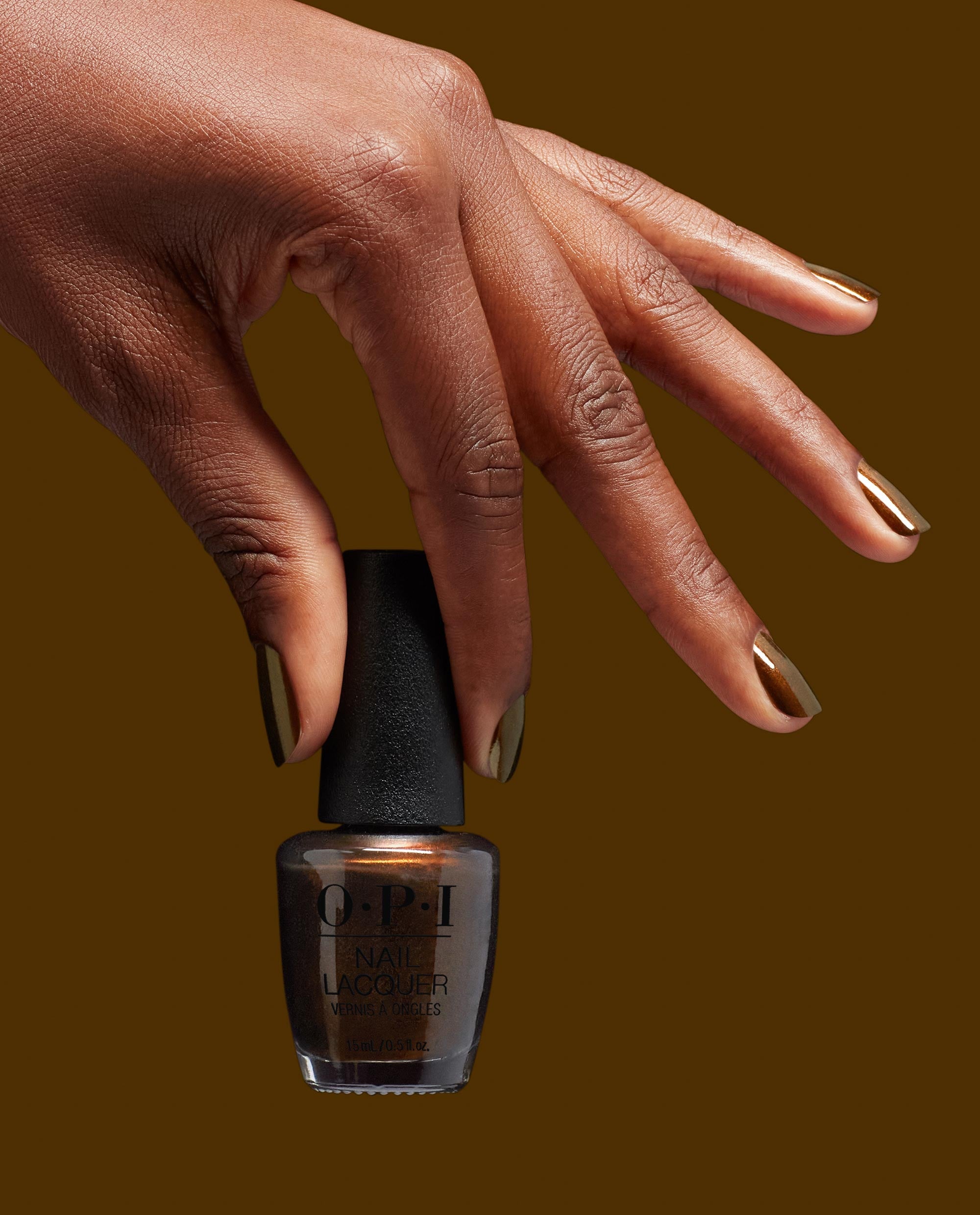 Hand with metallic bronze Band Tease OPI nail polish on neatly manicured nails gracefully holding the Band Tease nail lacquer bottle against a rich brown background, showcasing the shimmery OPI nail polish shade for US beauty shoppers.