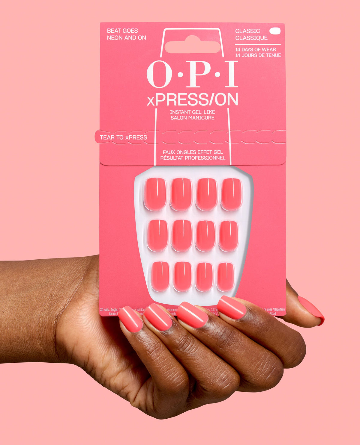 Hand with glossy coral-pink press-on nails holds OPI xPRESS/ON Beat Goes Neon and On artificial nail pack, showing matching gel-like nails on a pink background for a trending manicure.