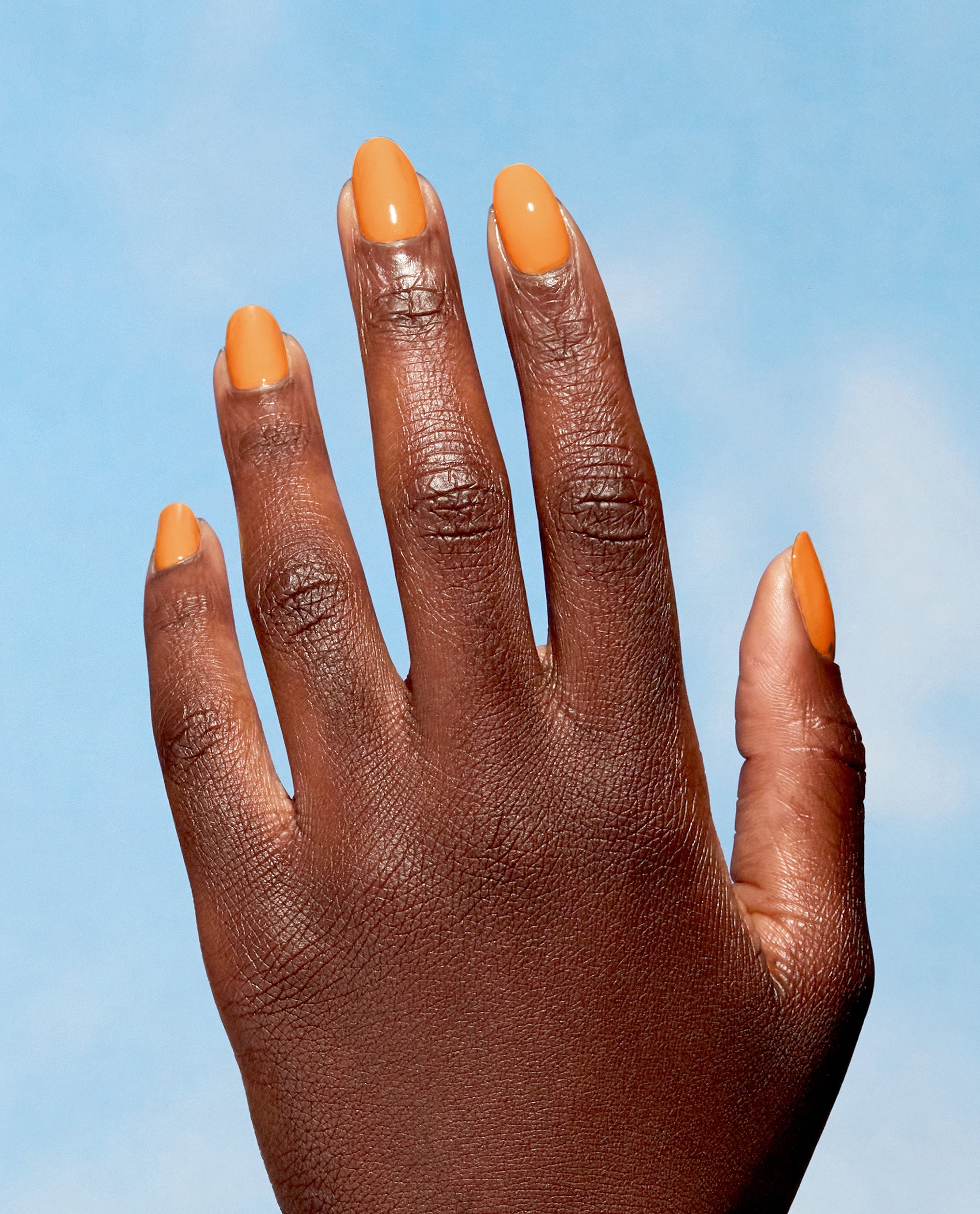 Hand showcases glossy Bee the Change manicure, a sunny marigold‑orange creme from OPI Nature Strong Vegan Nail Lacquer, shining against a clear blue sky - perfect for spring nail colors and spring nail trends from OPI.