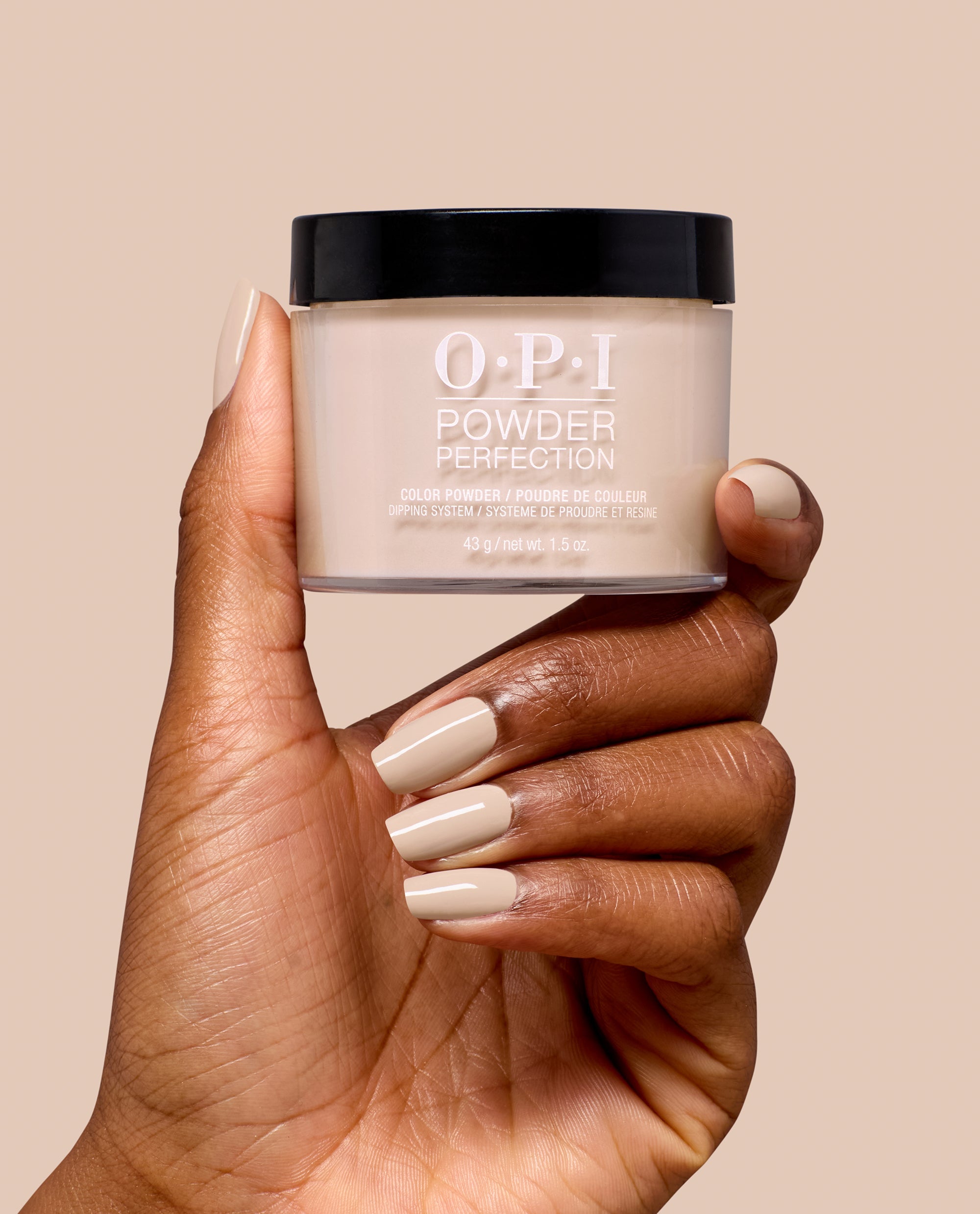 Hand with glossy beige taupe manicure holds OPI Powder Perfection Berlin There, Doin’ That dipping powder jar, a neutral shade for classic fall nail colors.
