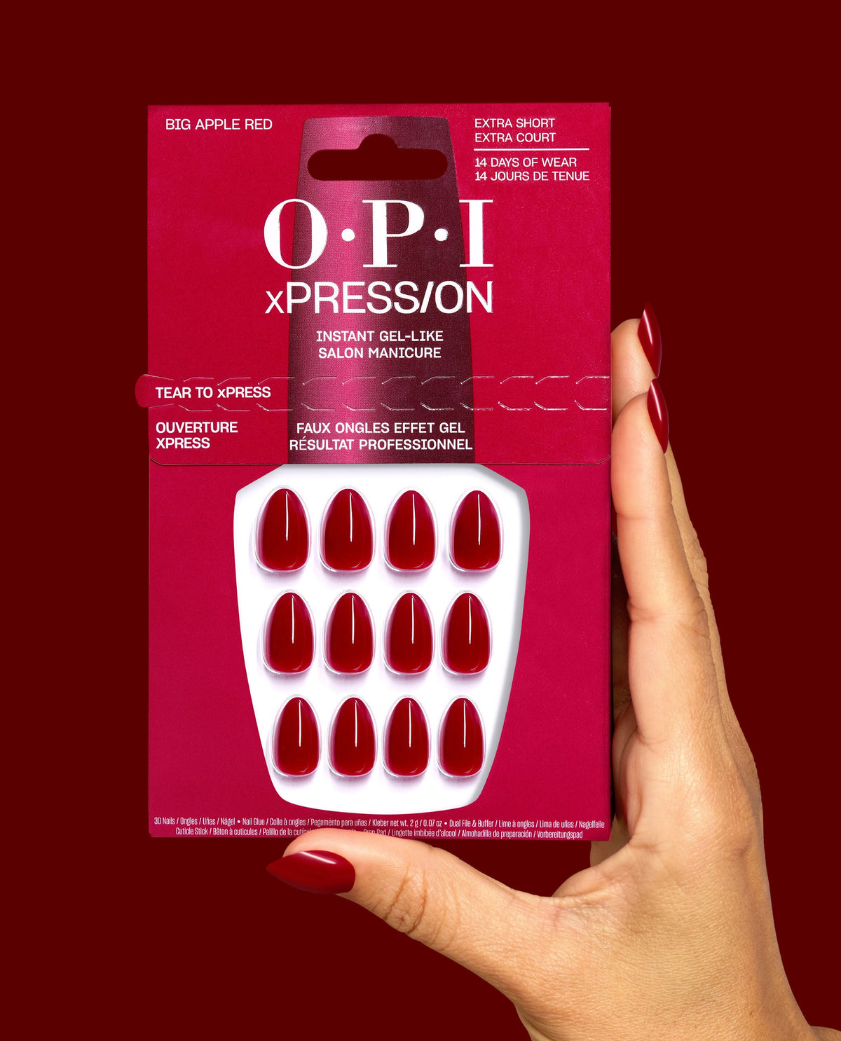 OPI xPRESS/ON Big Apple Red extra short press-on nails are held in hand, showing glossy deep red gel-like nails and matching manicure against a dark red background, ideal for fall nail colors.