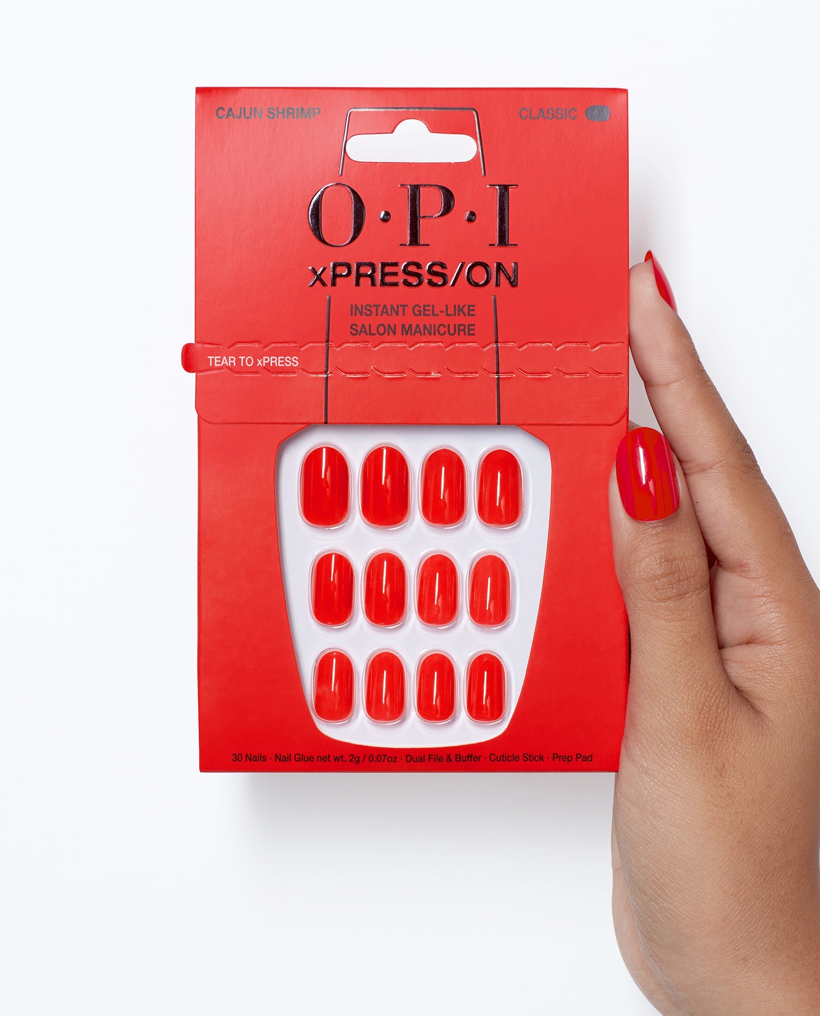 OPI xPRESS/ON Cajun Shrimp press-on nails in glossy coral red are shown in red packaging, held by a hand wearing the trending shade against a clean white background.